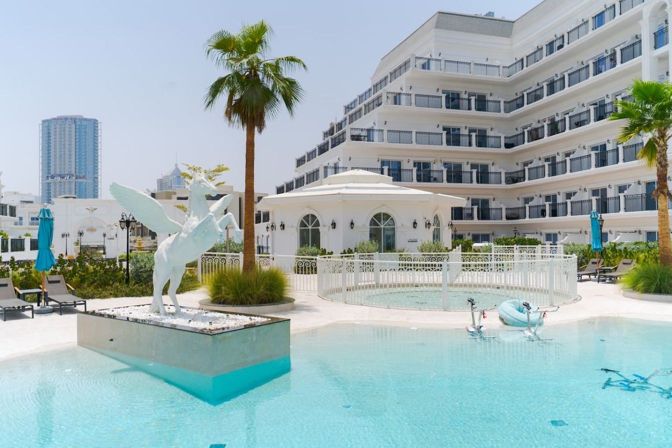 Zona de piscina de hotel con un edificio blanco, palmeras y una estatua.