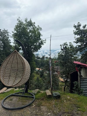 A wicker chair, a metal pole, and a red-roofed structure in a natural setting with trees and a distant view. - New Hotel Sajwan (Ukhimath)