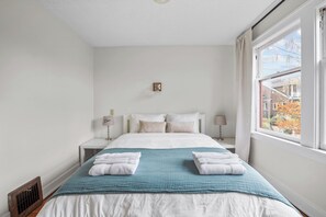 Une chambre à coucher avec un lit, deux tables de chevet, une fenêtre et une vue sur les bâtiments environnants.