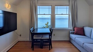 Une pièce comprenant une table à manger et des chaises, un canapé, un téléviseur fixé au mur et une fenêtre donnant sur un plan d’eau et des montagnes.