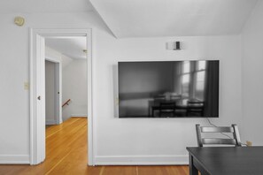 A room with a flat-screen TV mounted on the wall, a dining table with chairs, and a wooden floor.