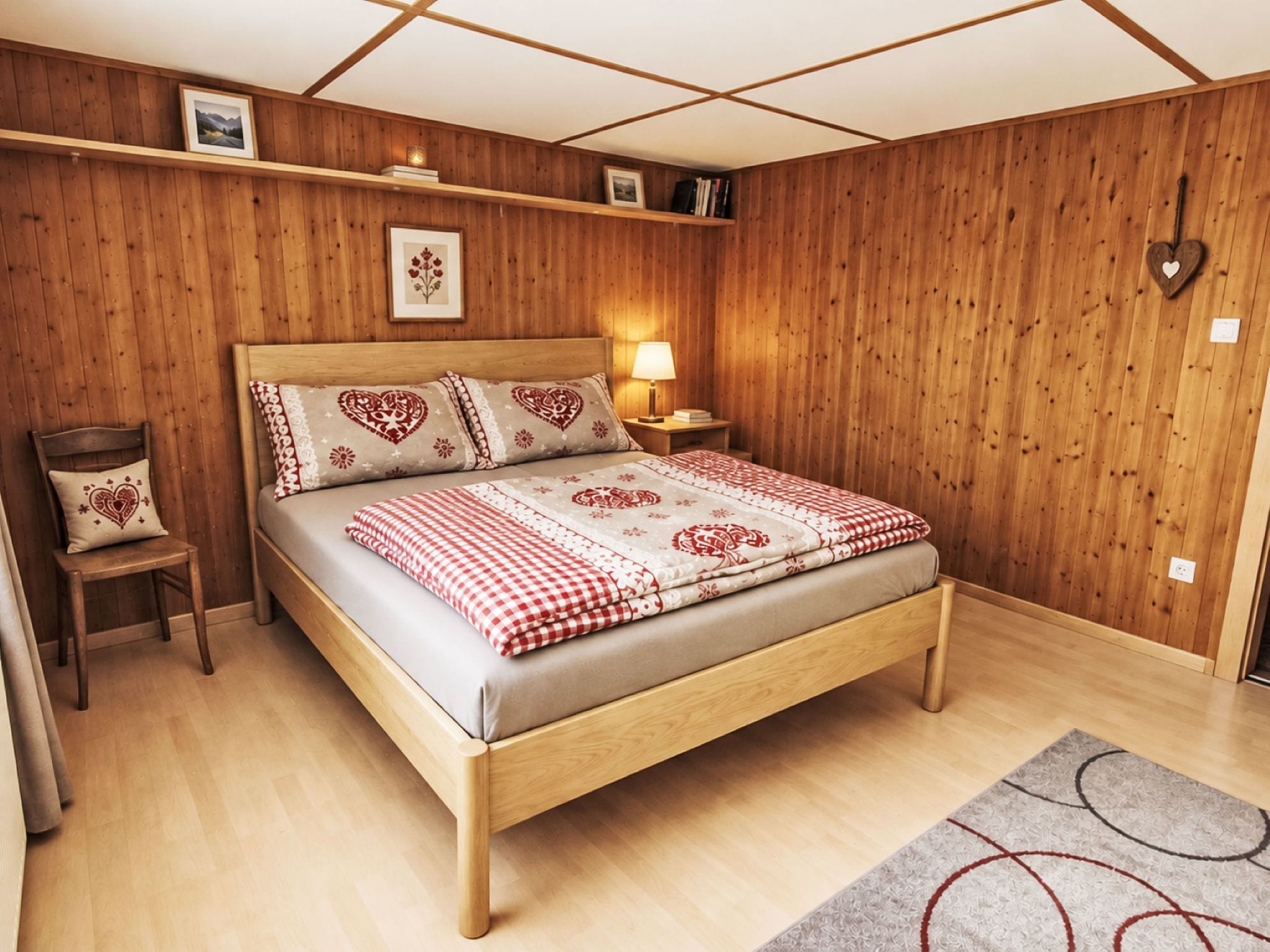 A wooden-paneled bedroom with a bed, a nightstand, a chair, and a wall-mounted heart-shaped decoration.