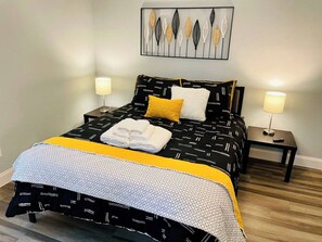 Een bed met een geborduurd beddegoed, twee kussens, een opgevouwen handdoek en een nachtkastje met een lamp.