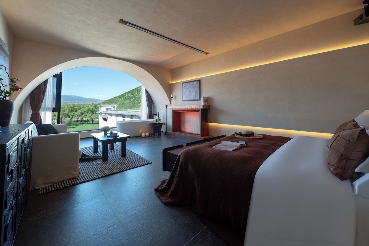 Habitación moderna con sofá, mesa de comedor y chimenea. Se observa una montaña y un edificio a través de un ventanal arqueado.