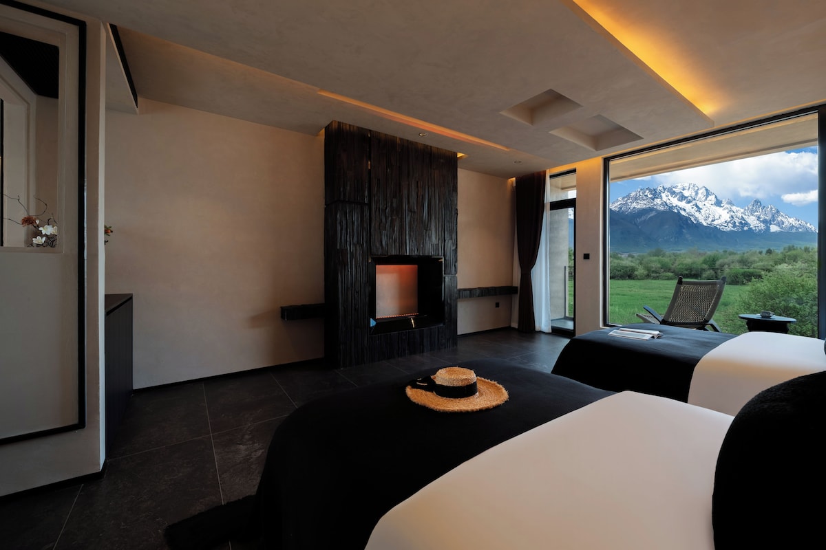 Una habitación de hotel moderna con chimenea, dos camas, vistas a montañas y un área de comedor con una mesa y sillas.