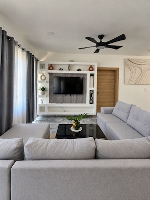 Ein modernes Wohnzimmer mit Flachbildfernseher, grauen Sofas, einem Couchtisch und einem Deckenventilator.