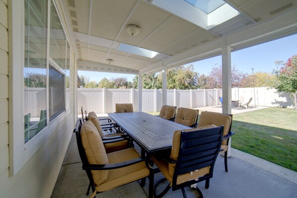 A covered patio with a table and chairs, a view of the backyard, and a white fence. - The Boise Retreat | Spacious Home, Game Room! (Boise)