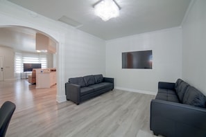 Ett modernt vardagsrum med en grå soffa, en platt-TV som är monterad på väggen och ett köksområde i bakgrunden.