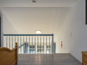 A spacious room with a wooden floor, a staircase with a blue handrail, and a view of an outdoor area. - Lütt Stuuv, Germany (Sylt)