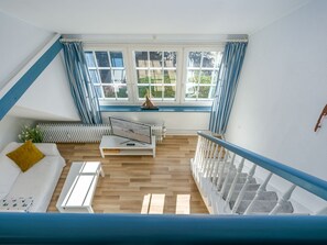 A room with a white sofa, a small table, a TV on a stand, a window with curtains, and a staircase with white balusters. - Lütt Stuuv, Germany (Sylt)