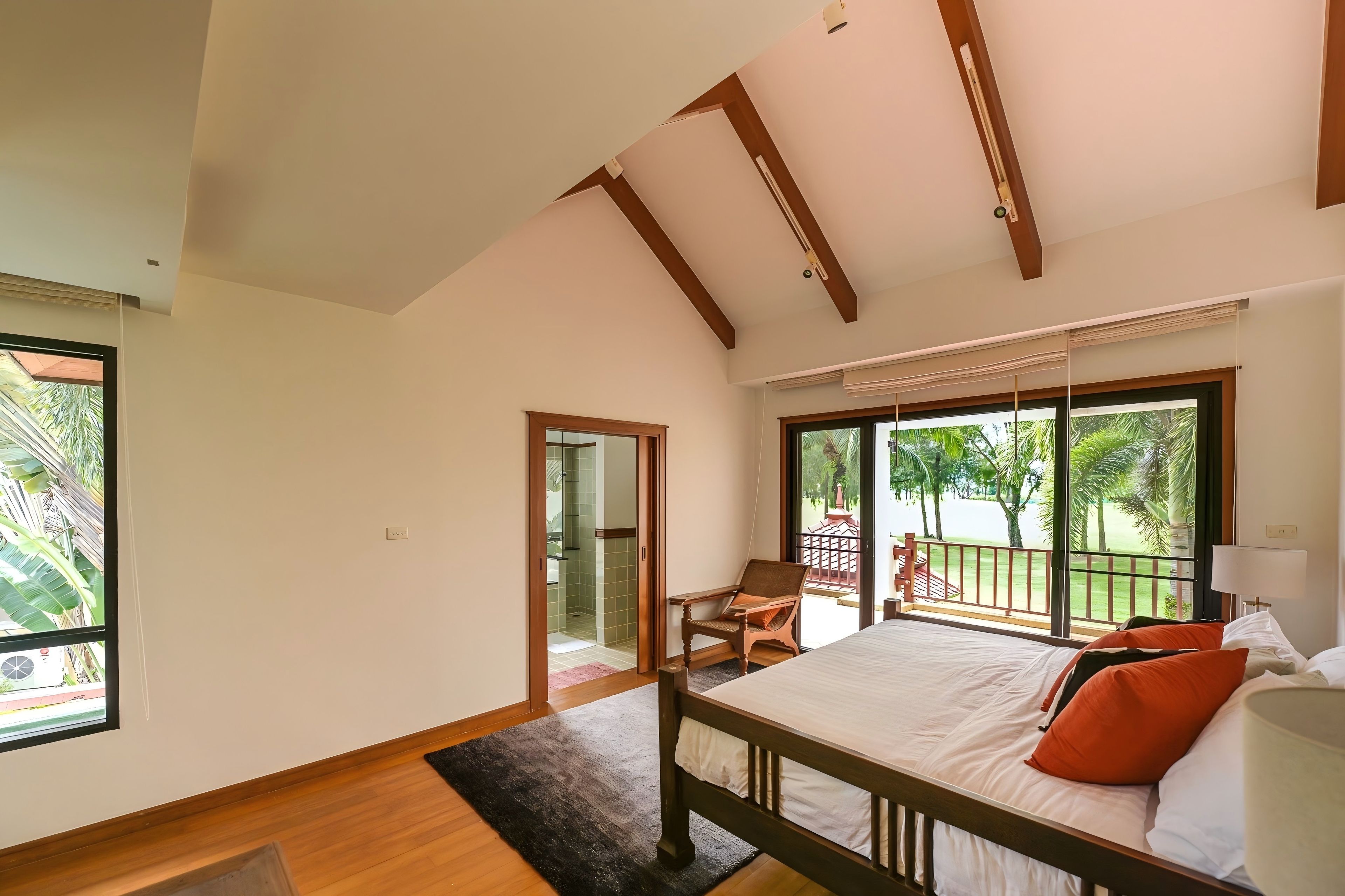 A bedroom with a large bed, a wooden headboard, a window with a view of greenery, and a sliding glass door leading to a balcony.