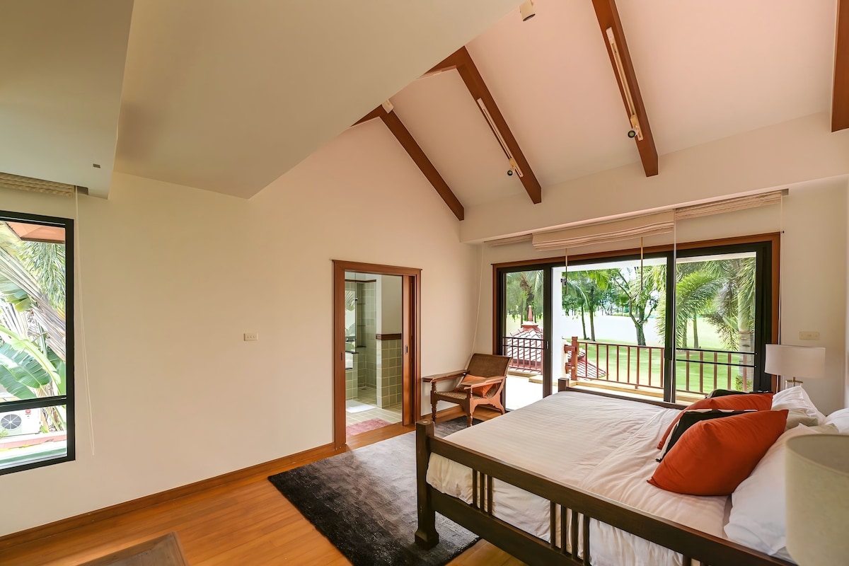 A bedroom with a large bed, a wooden headboard, a window with a view of greenery, and a sliding glass door leading to a balcony.