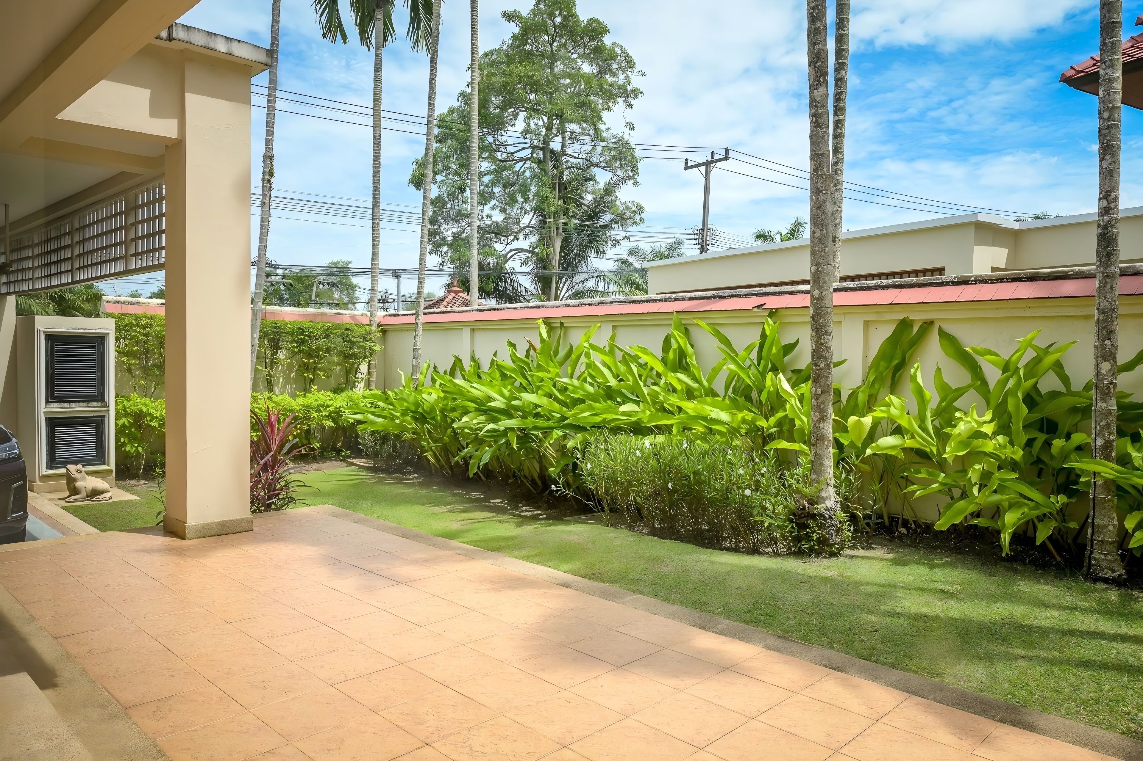 A residential area with a paved walkway, a row of green plants, and a building with a red roof.