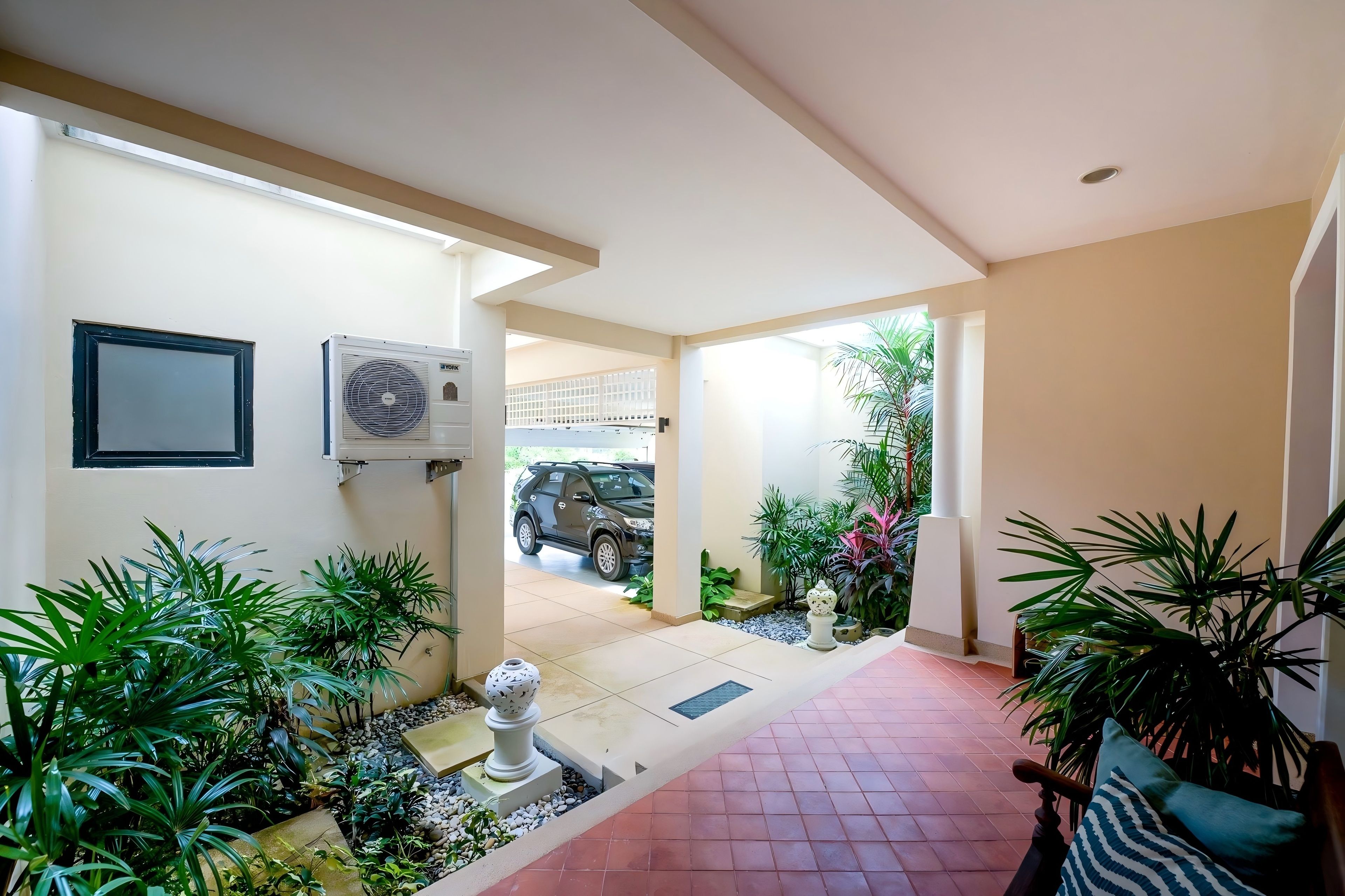 Un espace extérieur abrité où se trouve un véhicule garé au fond, un chemin menant à l’entrée d’un bâtiment, ainsi que diverses plantes et un vase décoratif.