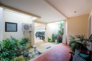 A covered outdoor area with a vehicle parked in the background, a pathway leading to a building entrance, and various plants and a decorative urn.