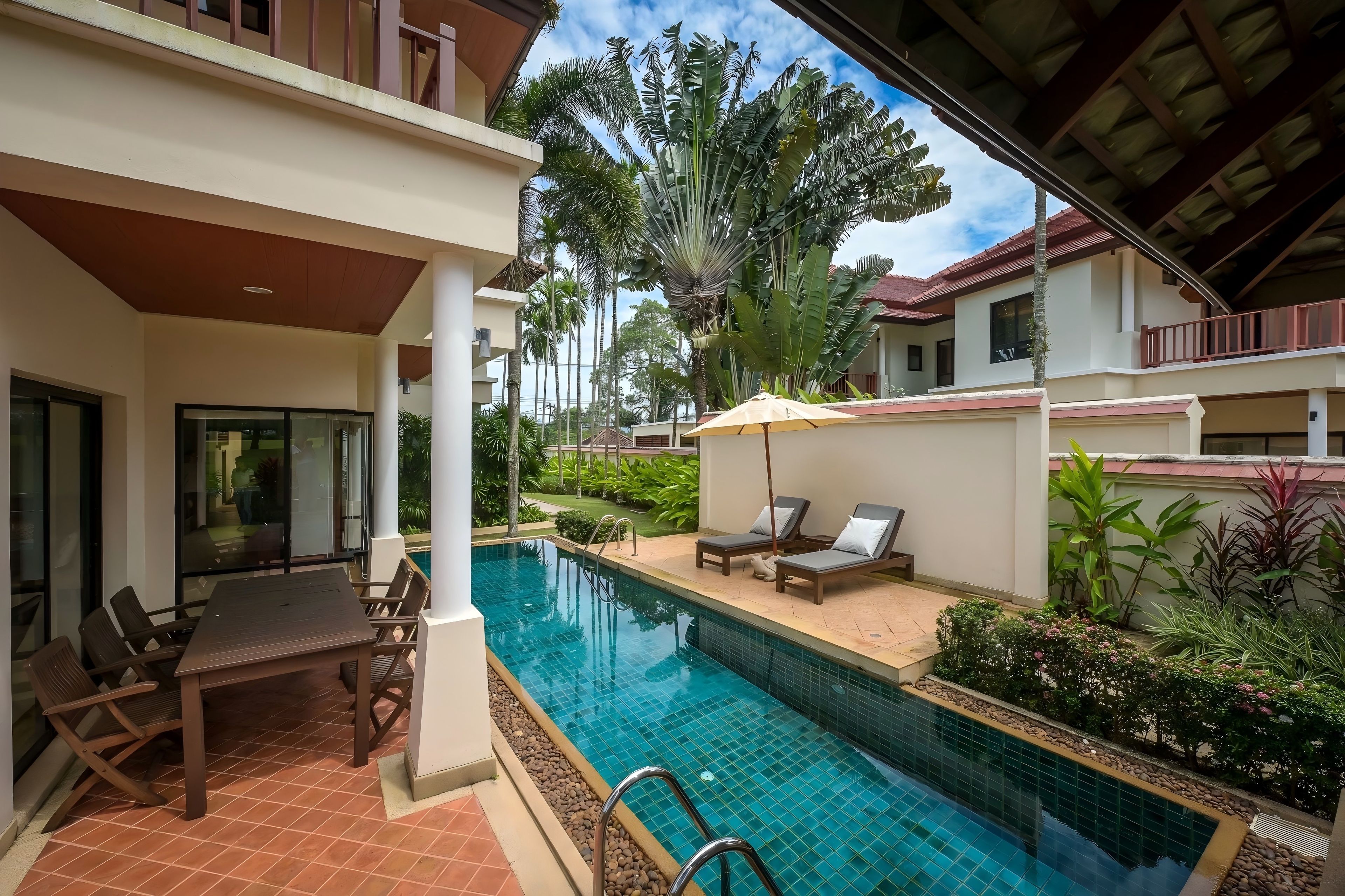 A swimming pool with lounge chairs and a table, surrounded by greenery and a covered patio area.