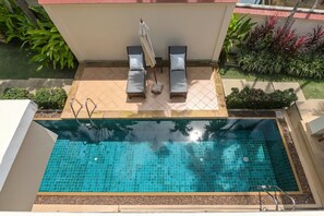 Una piscina con suelo de baldosas, dos sillas de descanso y un pequeño jardín con plantas.