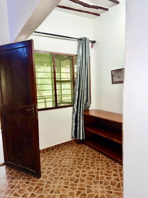 A room with a wooden door, a window with a metal screen, a grey curtain, a wooden bench, and a tiled floor. - MARUHUBI VILLAS (Zanzibar Town)