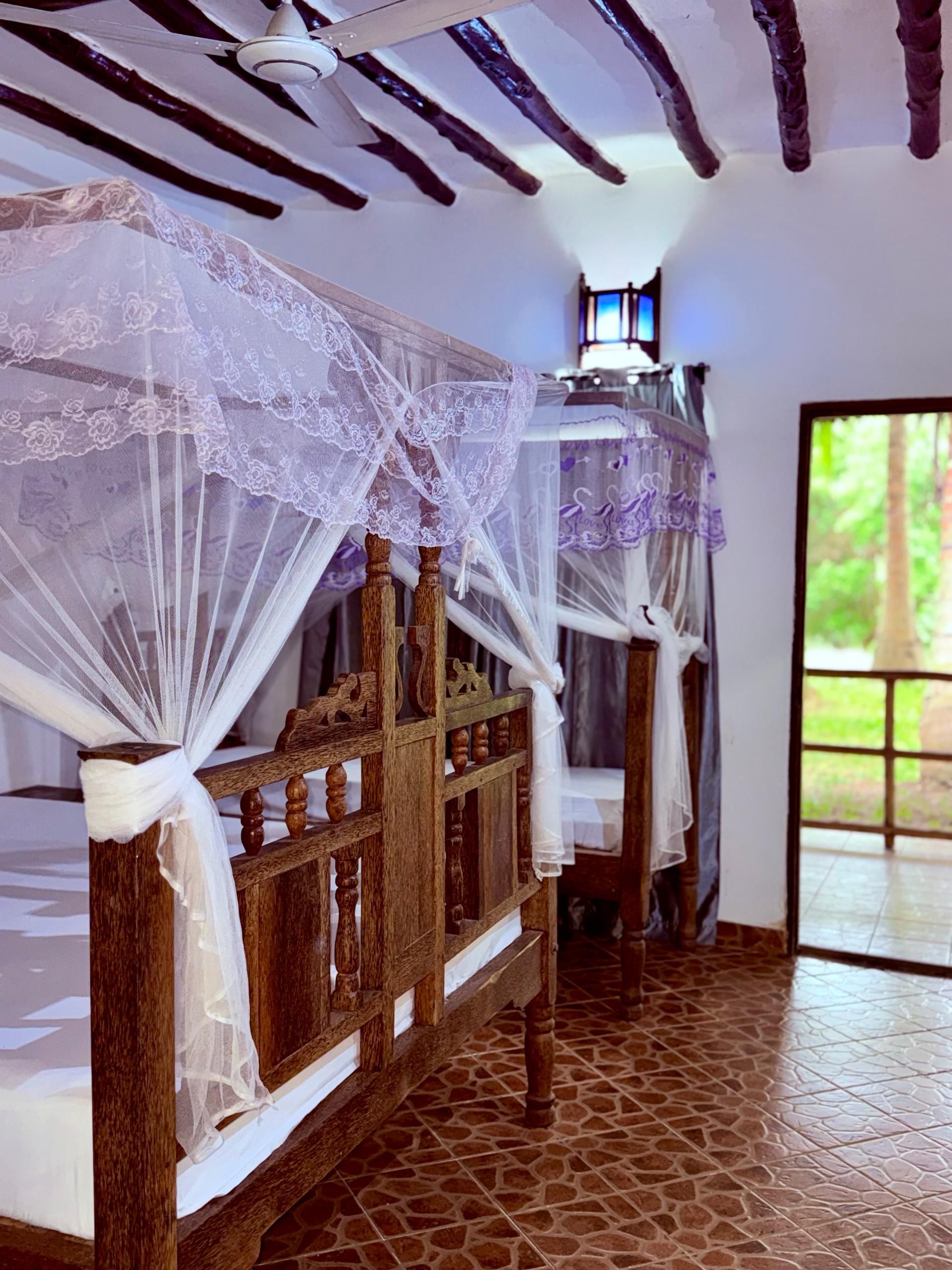 Una habitación con dos camas de madera, ropa blanca de cama con dosel de encaje, cabecero de madera y piso de baldosas.