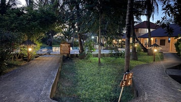 A well-lit outdoor area with a swimming pool, palm trees, and a paved walkway.
