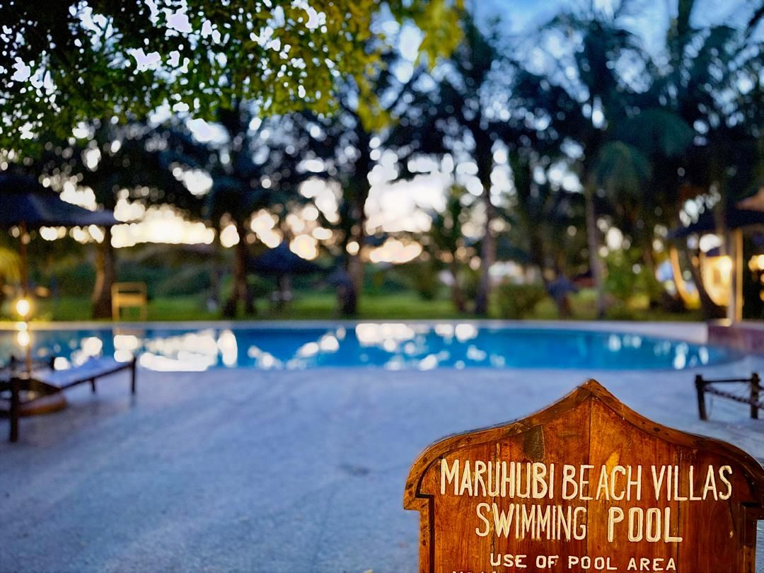 Un letrero de madera con el texto 'MARUHUBI BEACH VILLAS USO DE PISCINA ZONA DE PISCINA' en un entorno de resort.