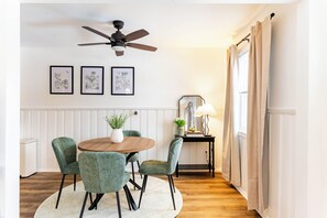 Un coin repas comprenant une table ronde, quatre chaises, un ventilateur de plafond et des reproductions encadrées de motifs botaniques accrochées au mur.