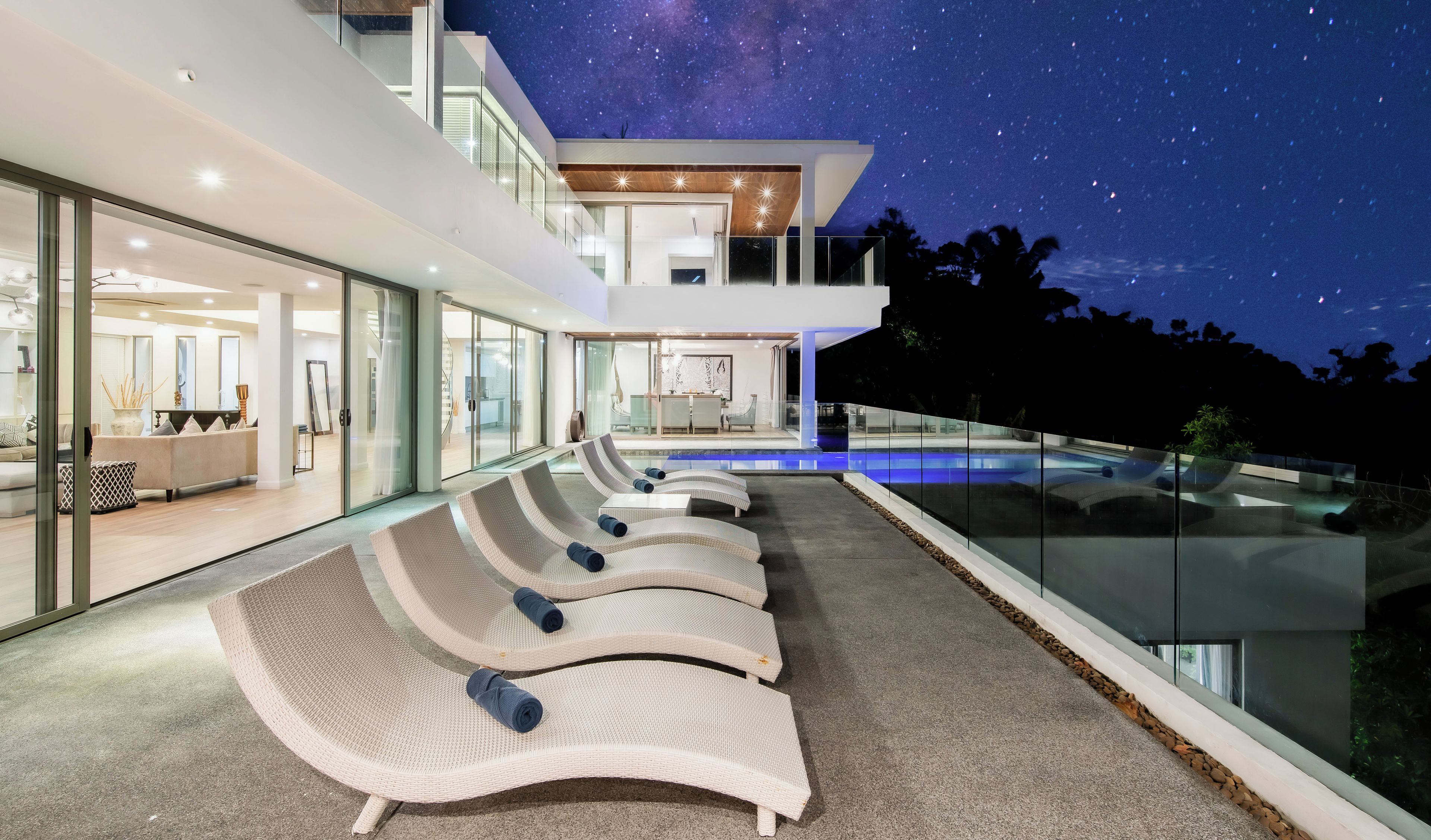 Una casa moderna con piscina, sillones de descanso y un cielo nocturno estrellado.