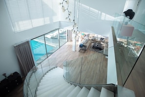 A modern interior with a glass staircase leading to a living area with a pool view. - MoonShadow Villa Phuket (Kathu District)