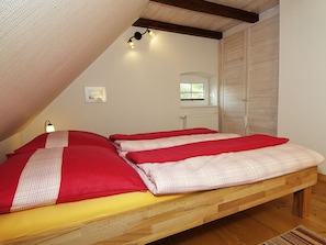 Ein Schlafzimmer mit schrägen Wänden, einem Bett mit rot-weißer Bettwäsche, einer Nachttischlampe, einem Fenster und einer Heizung.