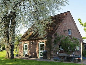 Ein Backsteinhaus mit grünen Türen und Fenstern, umgeben von einem Garten mit einem großen Baum und blühenden Blumen.