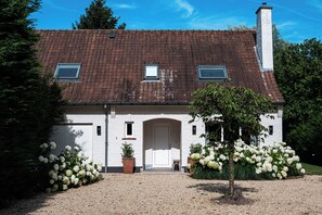 A two-story house with a tiled roof, a central entrance with a door, and a small garden with white flowers. - New Listing (Deurle)