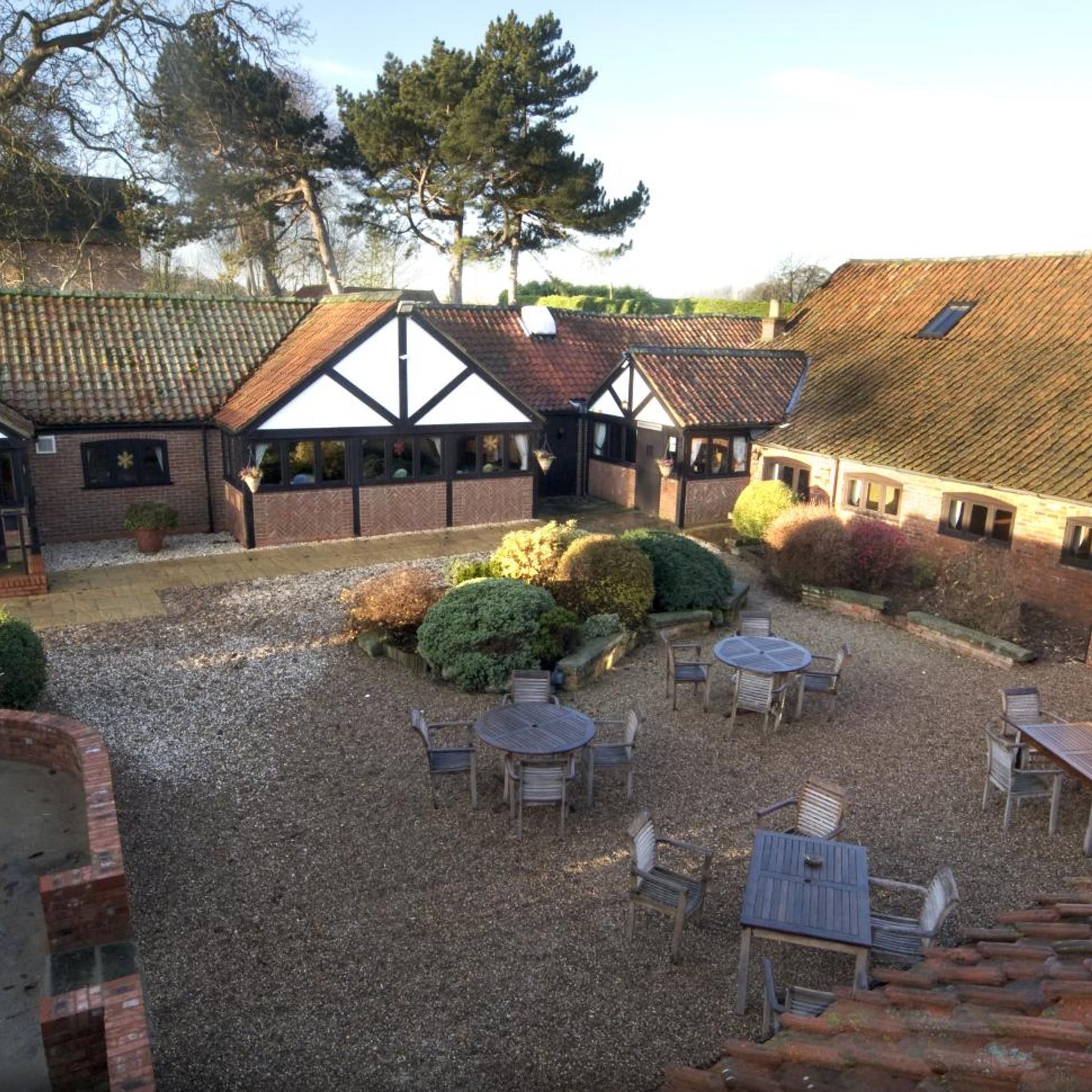 Un patio con suelo de grava, rodeado de edificios de arquitectura tradicional, incluyendo una estructura de estilo Tudor. Hay mesas y sillas al aire libre dispuestas para comer o socializar.