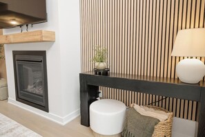 A modern living room with a fireplace, a black console table, a white ottoman, and a wall-mounted TV. - Muskoka Pines -Private Oasis Spa- Hot Tub & Sauna (Lake of Bays)