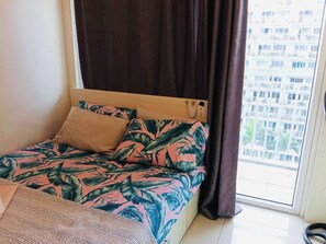 A bed with a floral bedspread, a wooden headboard, and a window with curtains. - Zen Escape 1Br with Balcony pacing pool (Pasay City)