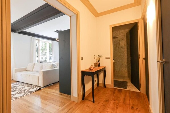 Una habitación con suelo de madera, un sofá blanco, una mesita con un florero y una puerta que da a un baño.