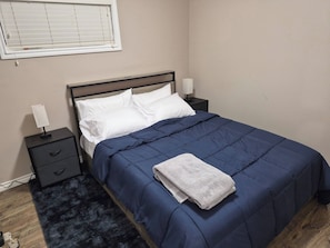 A bedroom with a bed, two bedside tables, a nightstand, a lamp, a window with blinds, and a dark blue rug. - D-King's Royal Court (Regina)