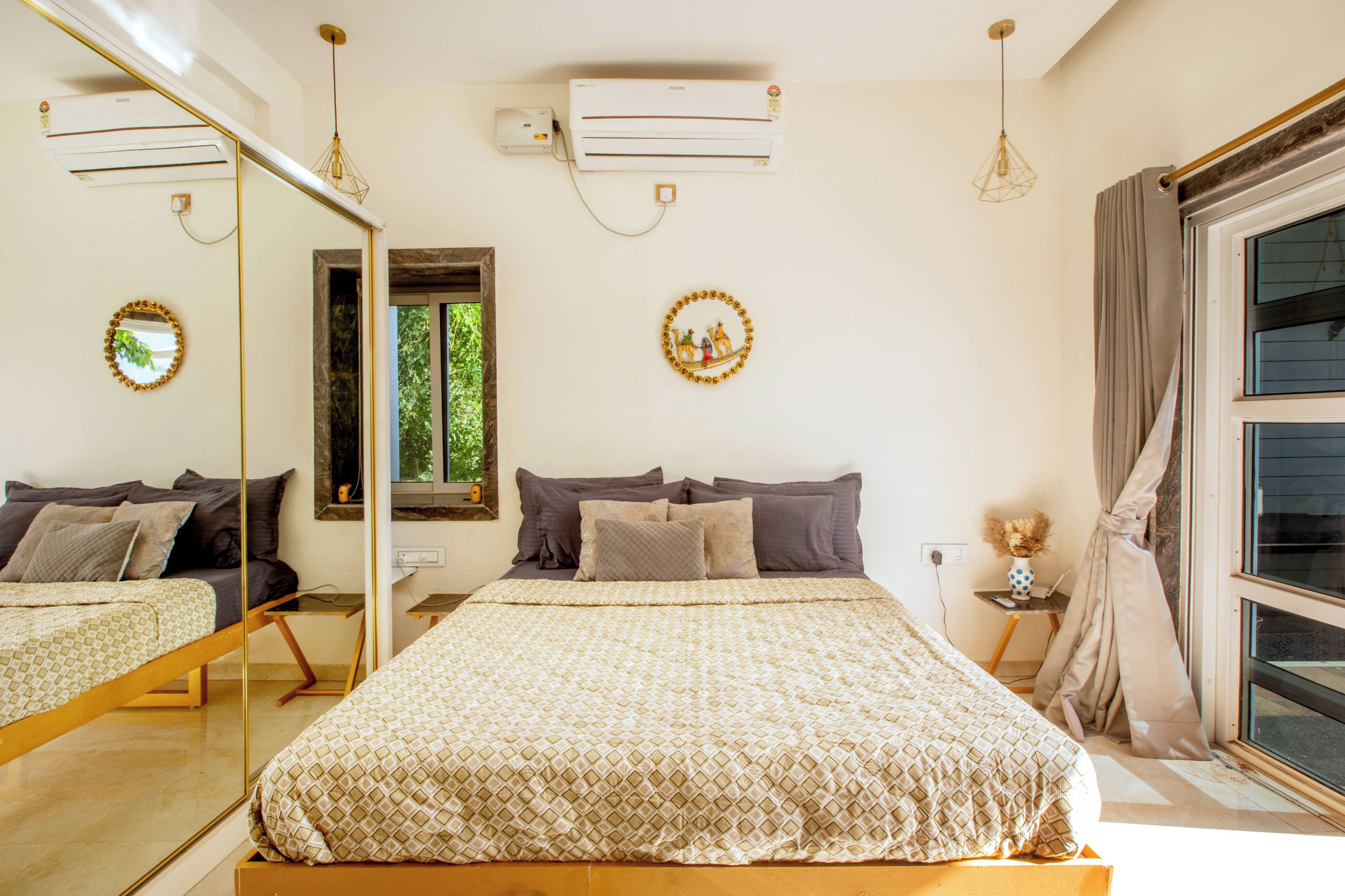 Un dormitorio con cama, espejo, equipo de aire acondicionado, ventana y mesita de noche con lámpara.