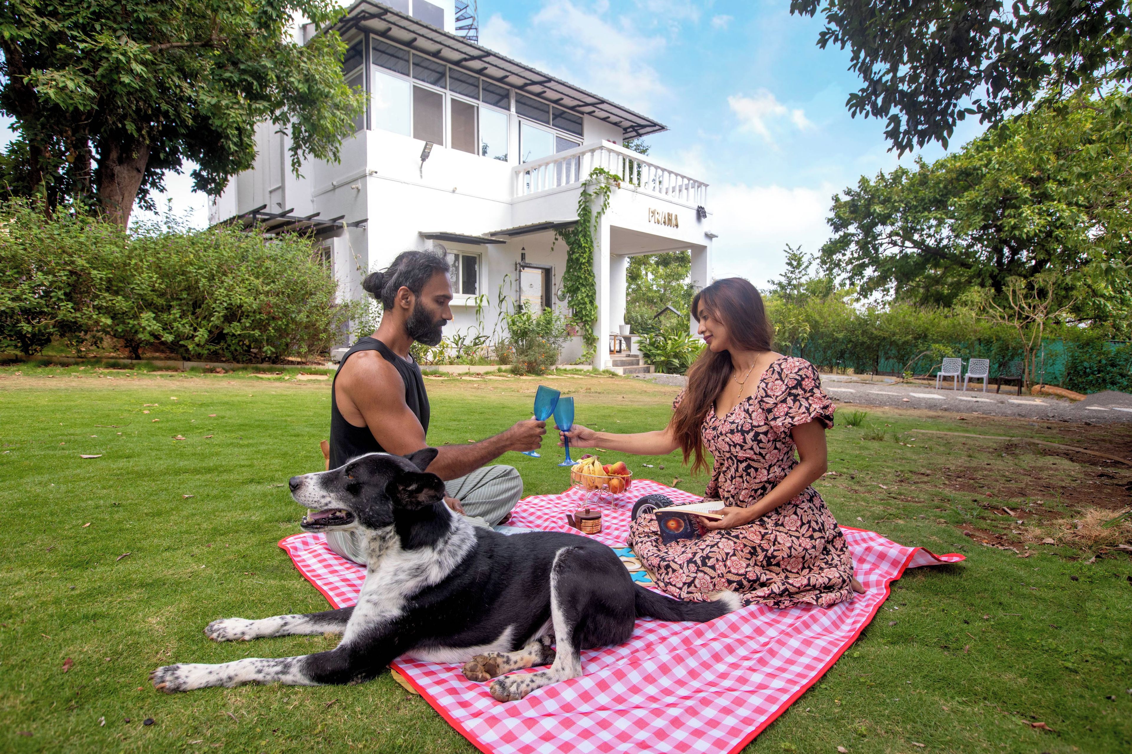 Dos personas sobre una manta a cuadros, un perro, una casa al fondo y cielo despejado.