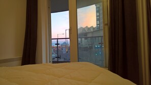 Une chambre d’hôtel avec un lit, une fenêtre avec des rideaux et une vue sur un paysage urbain composé d’immeubles et d’un coucher de soleil.