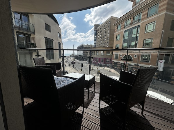 Ein Balkon mit Korbmöbeln, Blick auf eine Stadtstraße und Gebäude mit großen Fenstern.