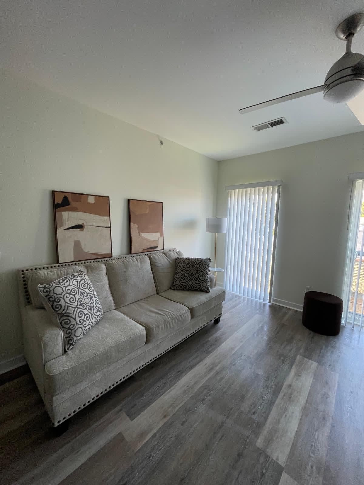 A modern living room with a beige sofa, two patterned cushions, a ceiling fan, and a large window with blinds.