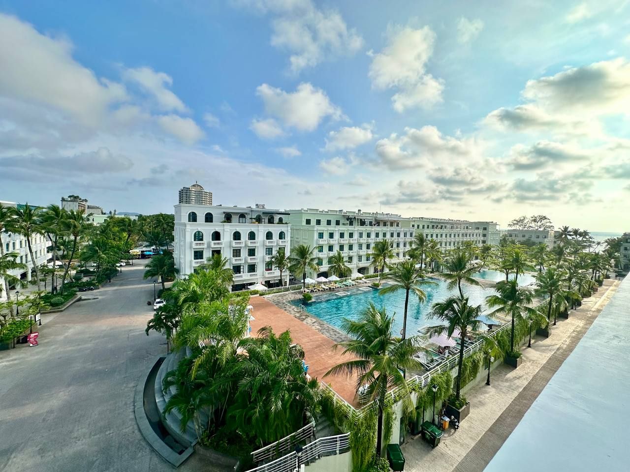 A hotel with a large pool surrounded by palm trees, a beach, and a clear sky.