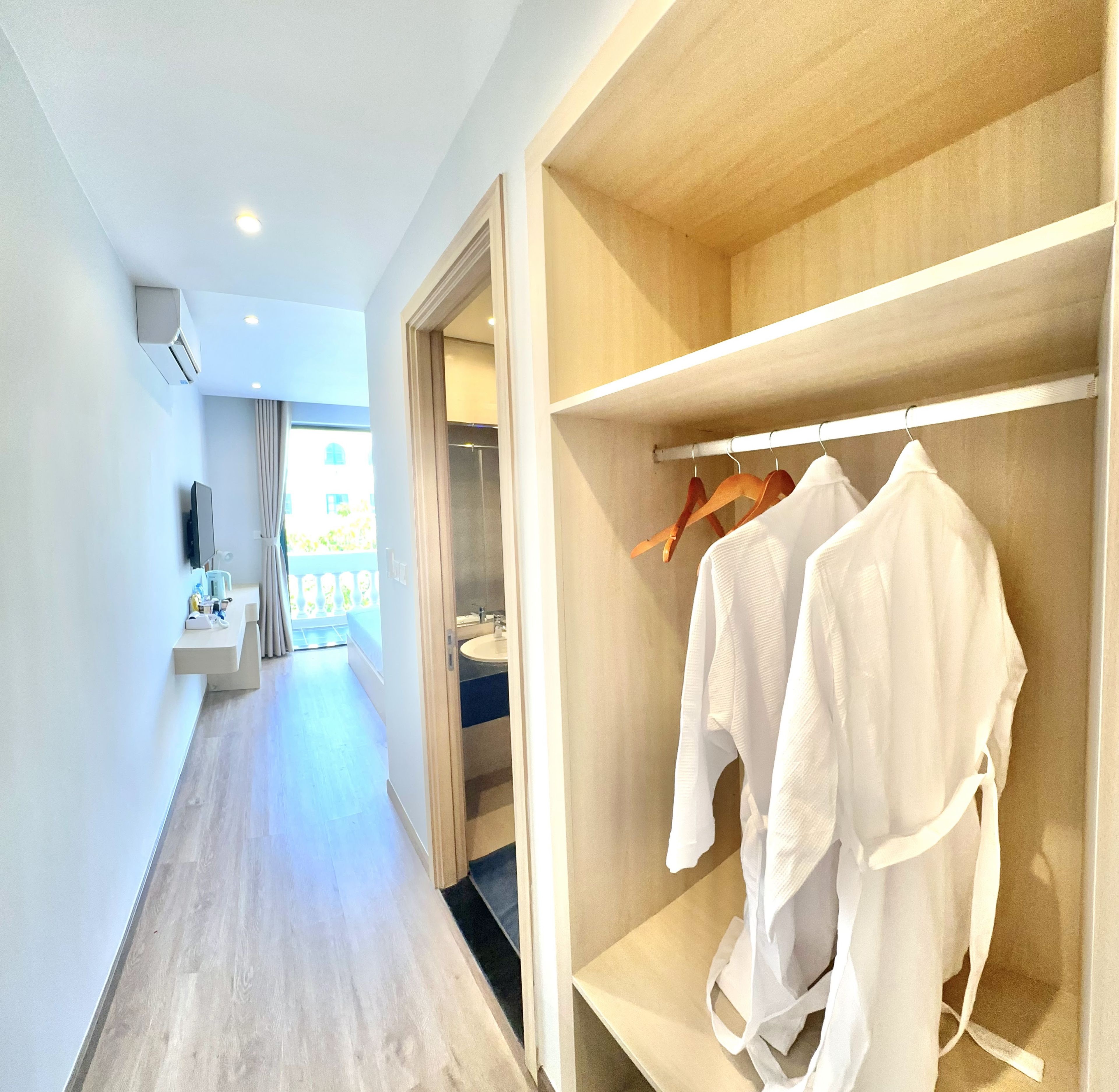 A hotel room with a wooden floor, a walk-in closet with white robes, a bathroom with a sink, and a view of a balcony.
