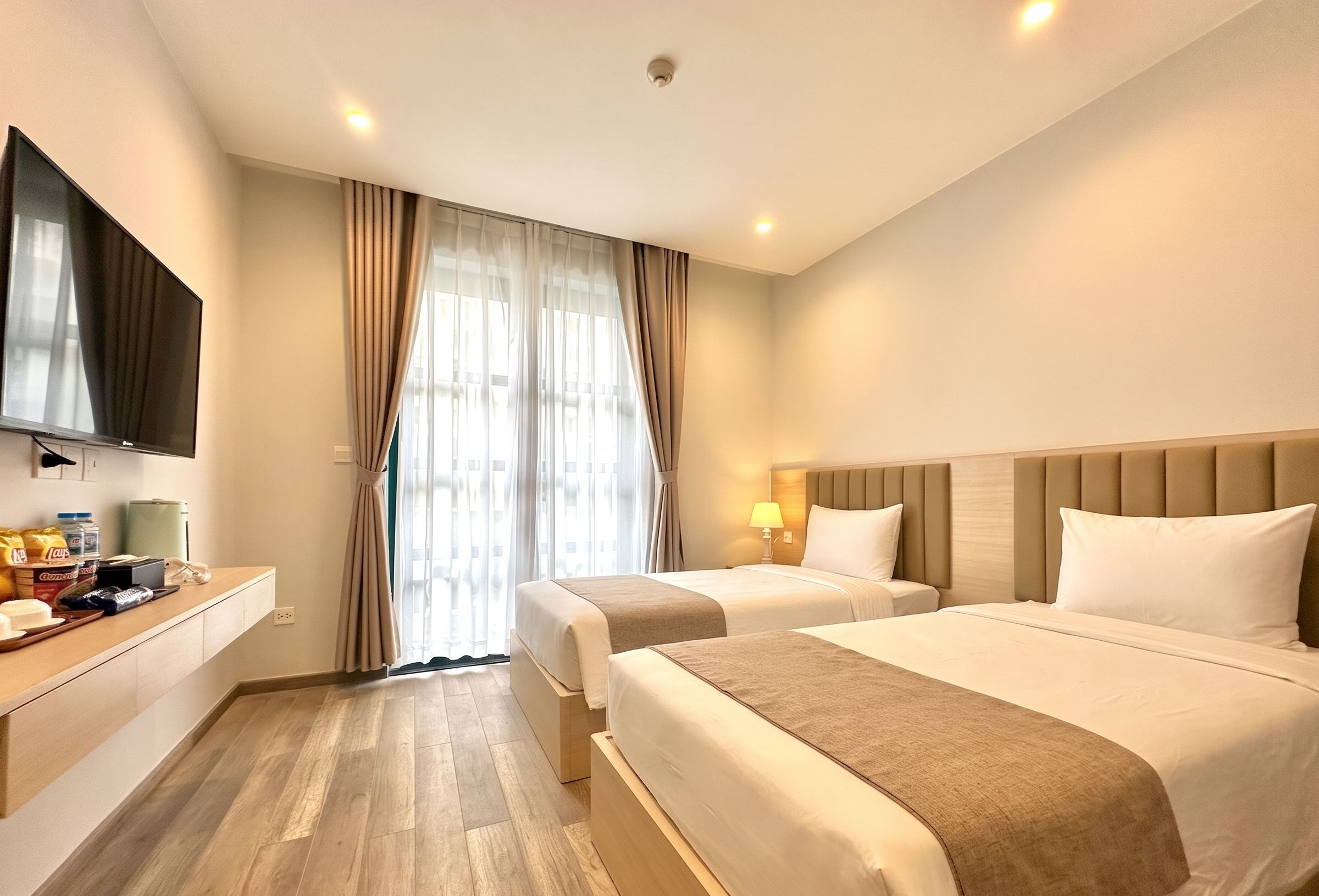 A hotel room with two beds, a TV, a bedside table, a lamp, and a window with curtains.