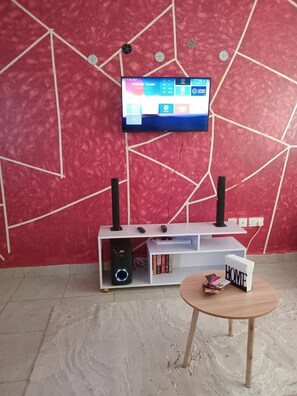 Ein Zimmer mit roter Wand, strukturierter Oberfläche, einem an der Wand befestigten Fernseher, einer weißen Unterhaltungseinheit mit Lautsprechern und einem kleinen runden Tisch mit einem Buch und einem Schild.
