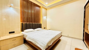A modern bedroom with a wooden headboard, a bed with white bedding, a nightstand, and a wooden wardrobe. - Hotel Rishant Palace (Omkareshwar)