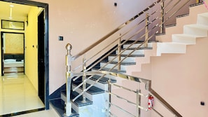 A staircase with a metallic handrail and black steps, leading to a room with a bed and a wall-mounted light fixture. - Hotel Rishant Palace (Khandwa)