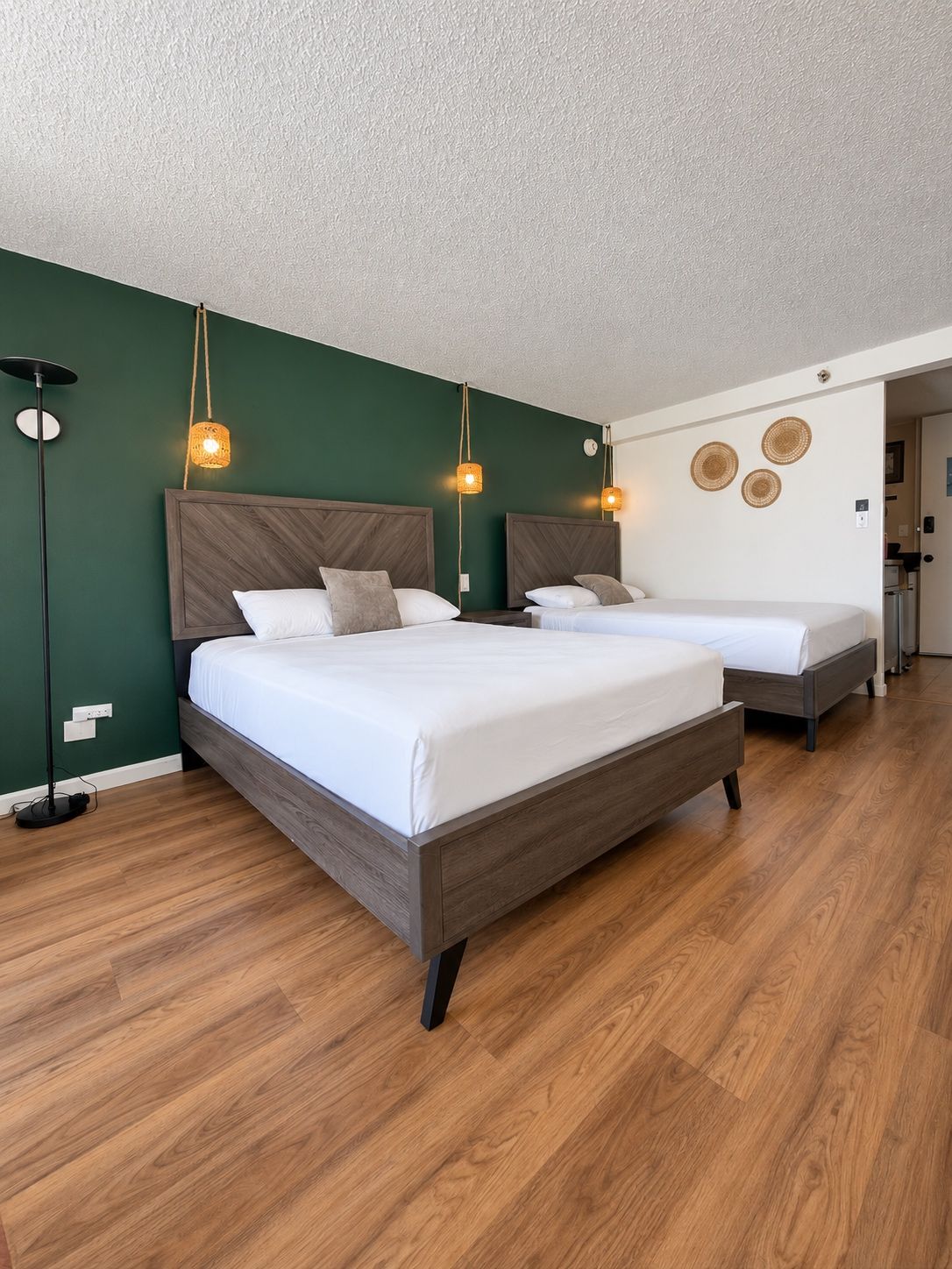 Habitación de hotel con dos camas, una pared verde, suelo de madera y una lámpara moderna.
