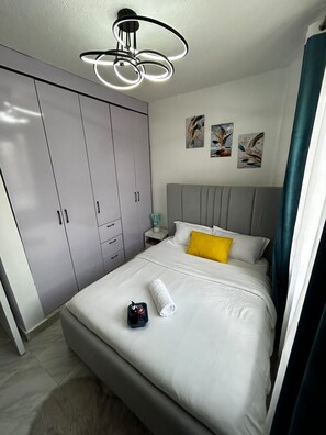 Ein Schlafzimmer mit einem Bett, einem grauen Wandschrank, Kunstwerken an der Wand und einer modernen Deckenleuchte.