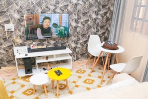 Ein Wohnzimmer mit Fernseher, weißen Stühlen, einem kleinen gelben Tisch und einer Wand mit Muster.